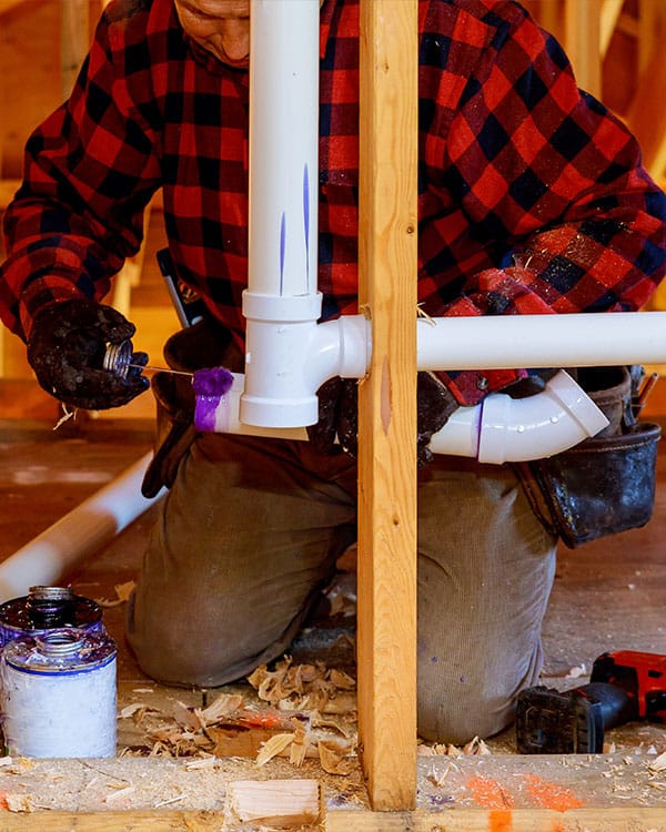 A plumber in flannel applies glue to a piece of pipe in a crawl space
